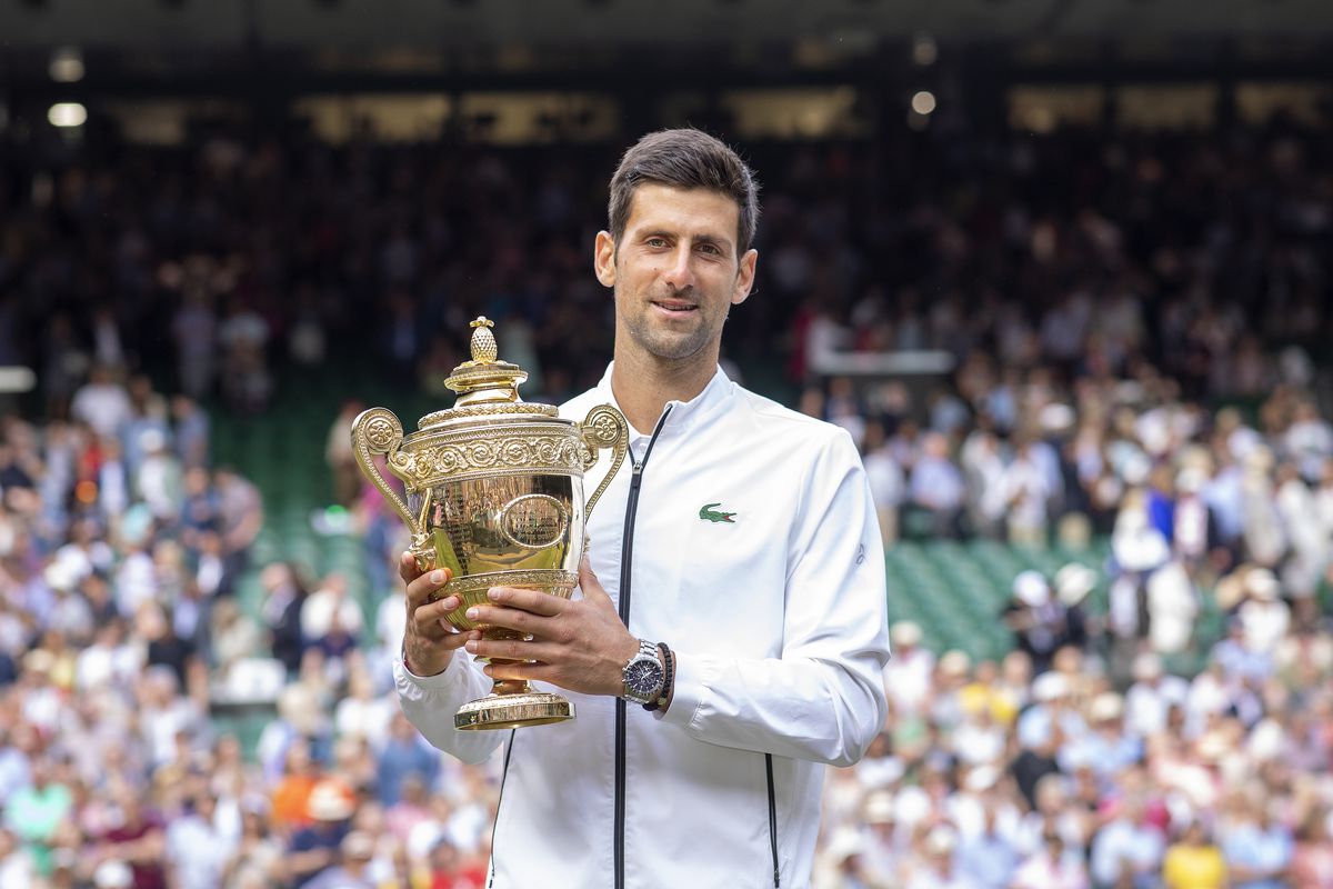 A man holds a golden trophy on a tennis court in England. A crowd of people is cheering behind him. 