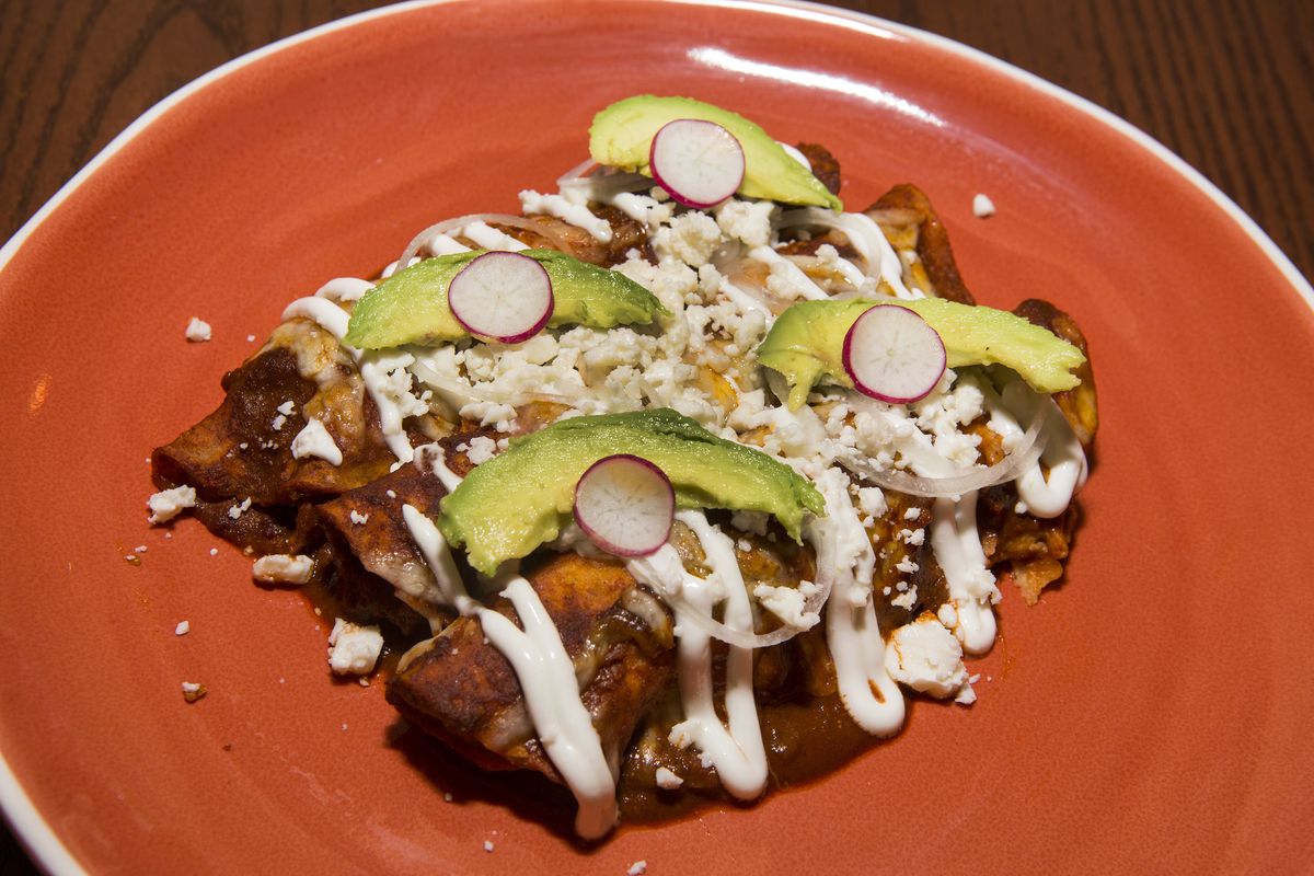 A red plate of enchiladas covered in a rich brown sauce with crema and cotija cheese. Slices of avocado and radish sit on top.