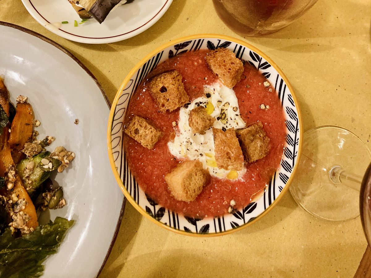 A bowl of bright red gazpacho with a blob of burrata in the middle and fried bread bits scattered around, on a table with other dishes and a pitcher of wine. 