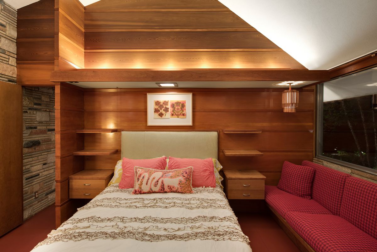 A small white bed with pink pillows sits in front of wood paneling and next to a red bench sofa. 