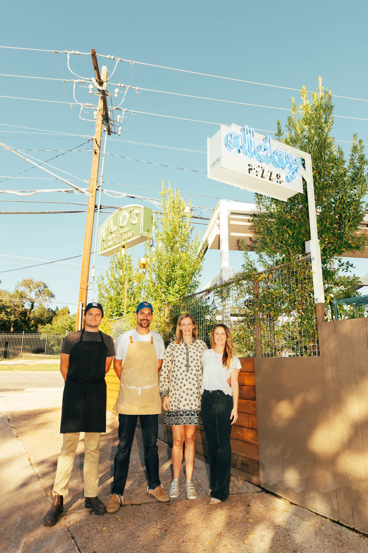 Four people posing underneath a sign outside reading Allday Pizza.