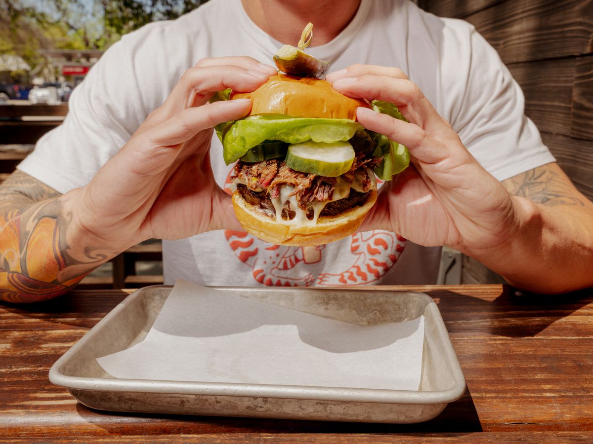 a person holding Loro burger that is bursting with beef patty, brisket, cheese, lettuce and thick-sliced pickles.