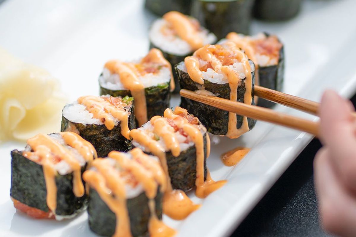 A sushi roll, drizzled in sauce, being picked up with chopsticks.