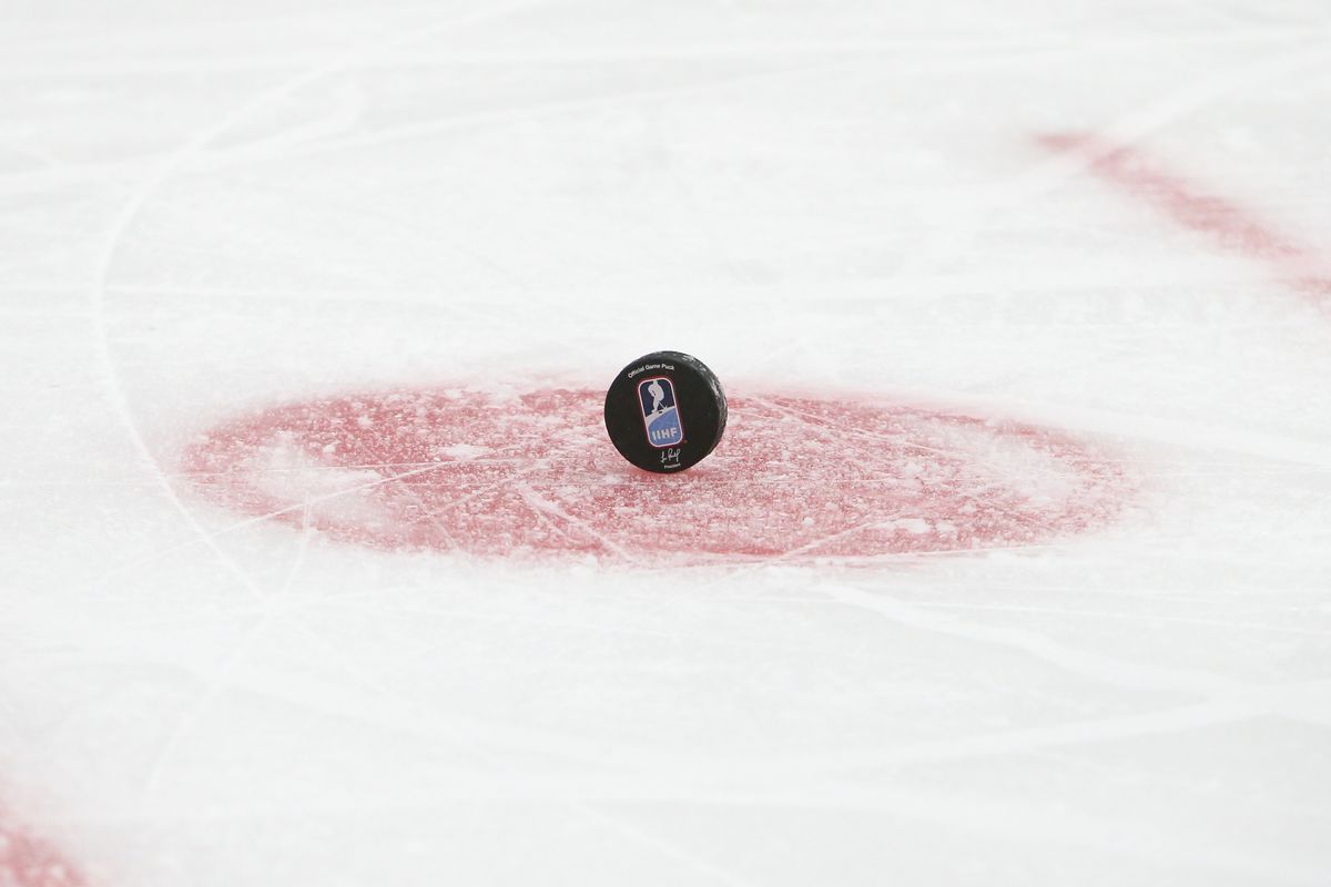 A black puck with the IIHF logo stands on its side on a red dot on white ice.