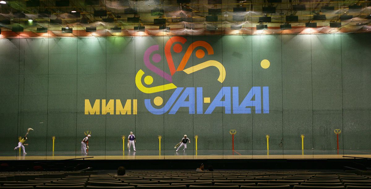 Players throw balls using hook shaped baskets on their arms in a large Jai alai court, with the words “Miami Jai Alai” artistically depicted on the background wall.
