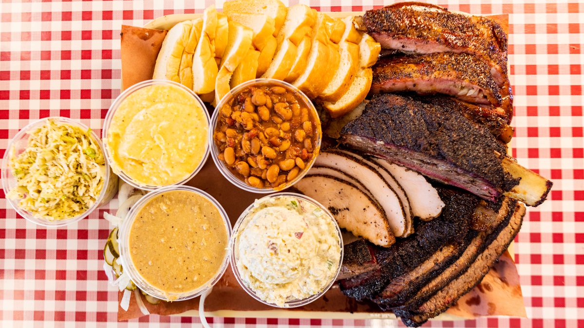 A platter of ribs, brisket, turkey, sliced bread, beans, potato salad, and other sides on a red and white checked tablecloth. 