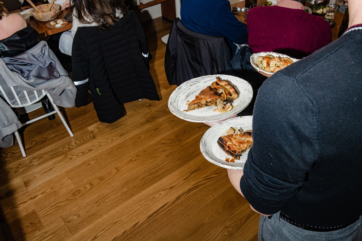 A person carrying three plates of food heads towards a table filled with diners in the background of the photo.