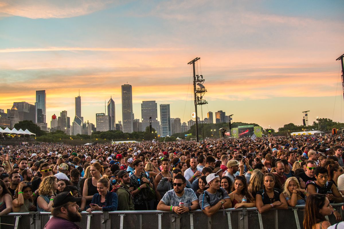 A Lollapalooza crowd looks at a massive stage behind the photographer. Sunset sky and Chicago skyscrapers in the back.
