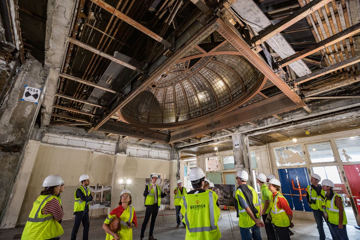A group of people in hardhats and yellow vests stand in the center of a gutted room. Above them is a large oval leaded-glass skylight.