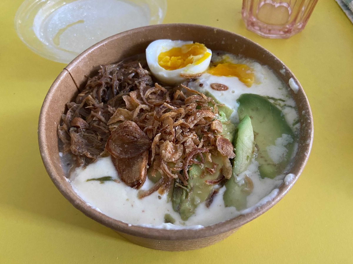 Congee with avocado, egg, and pork presented in a paper to-go bowl on a yellow table.