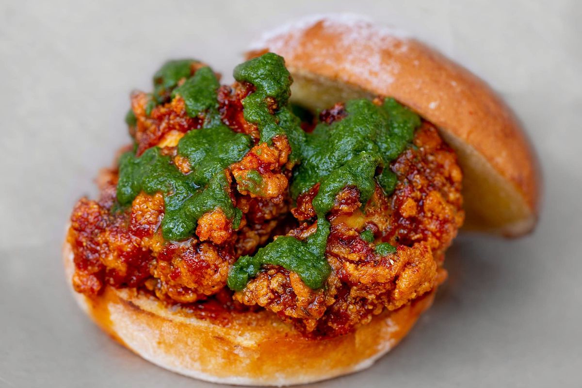 A fried chicken sandwich with the top bun propped aside to show the chicken drizzled in a green mint chutney.