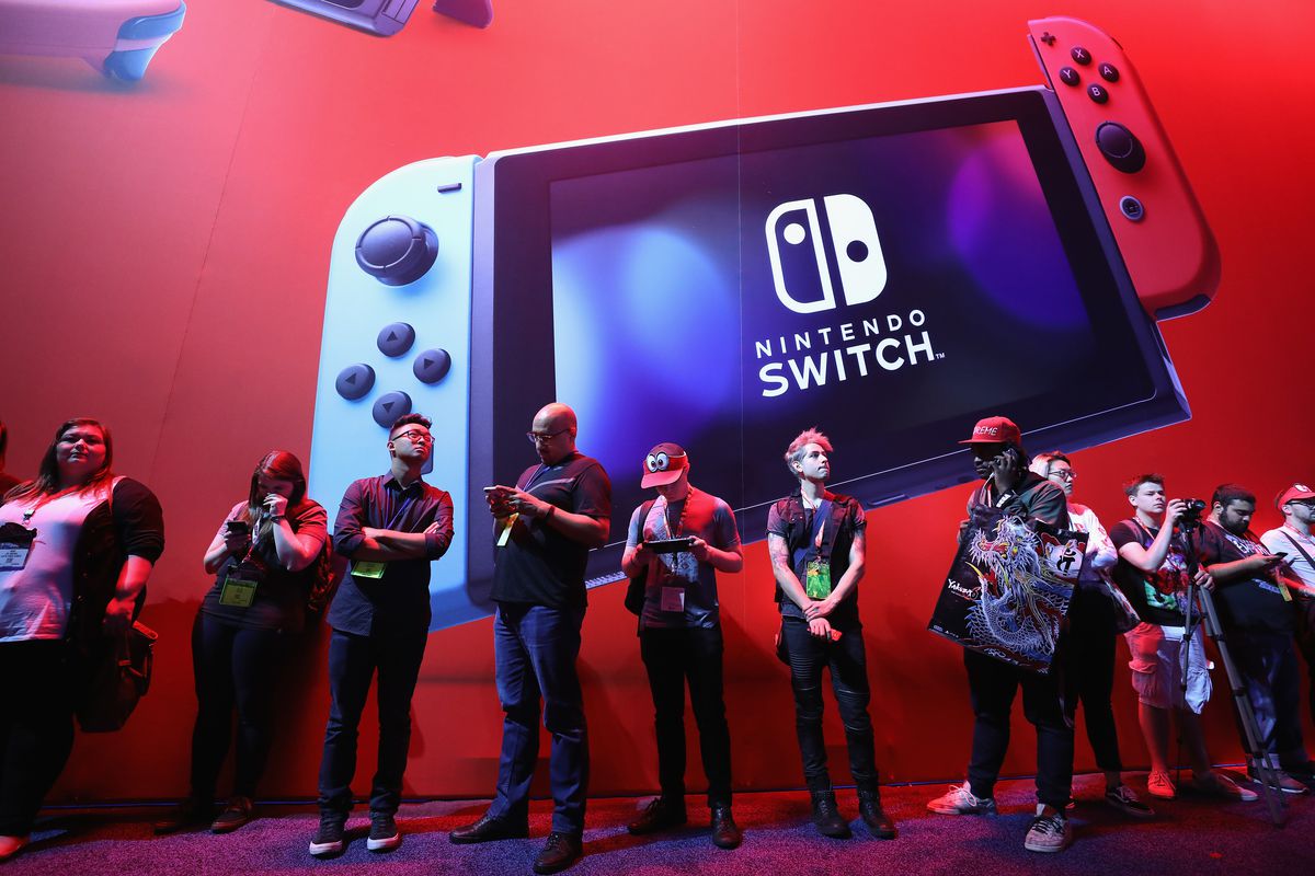 a bunch of people standing in line in front of a red wall with a large Nintendo Switch on it