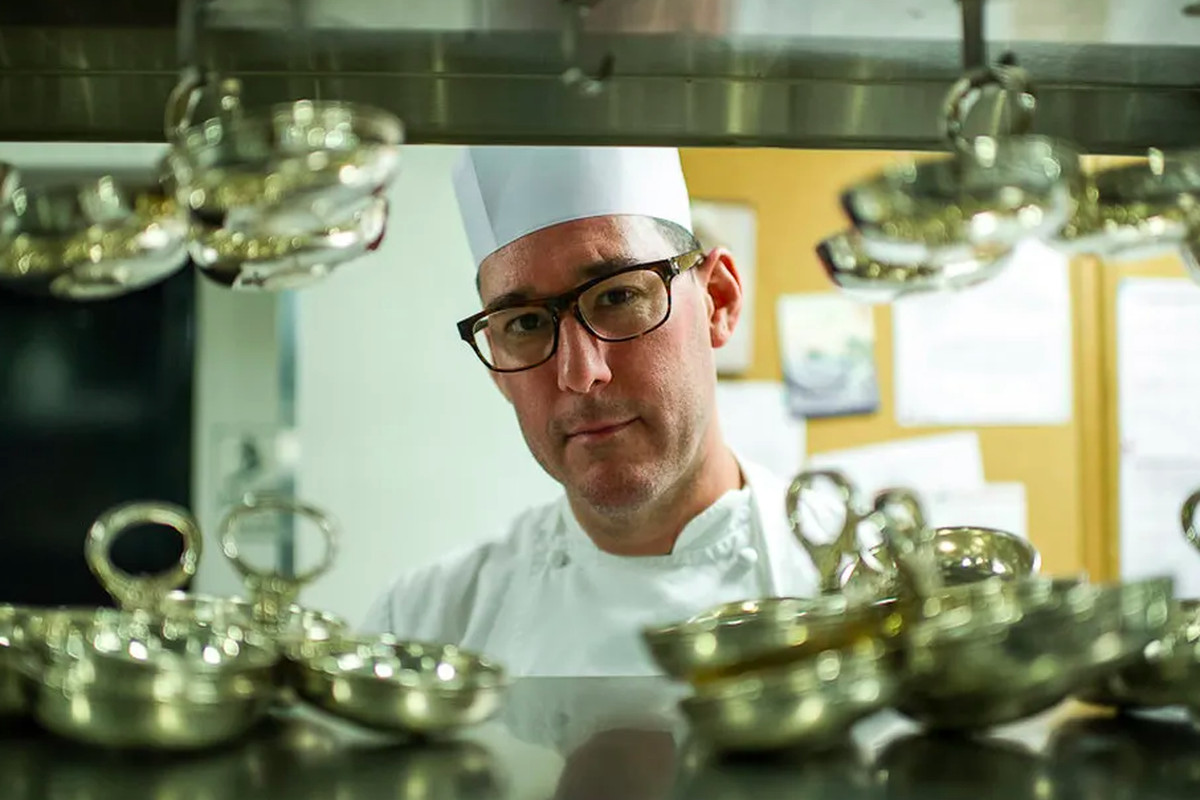 A white male chef wearing glasses with thick black rims stands behind the pass at a restaurant, wearing chef’s whites.