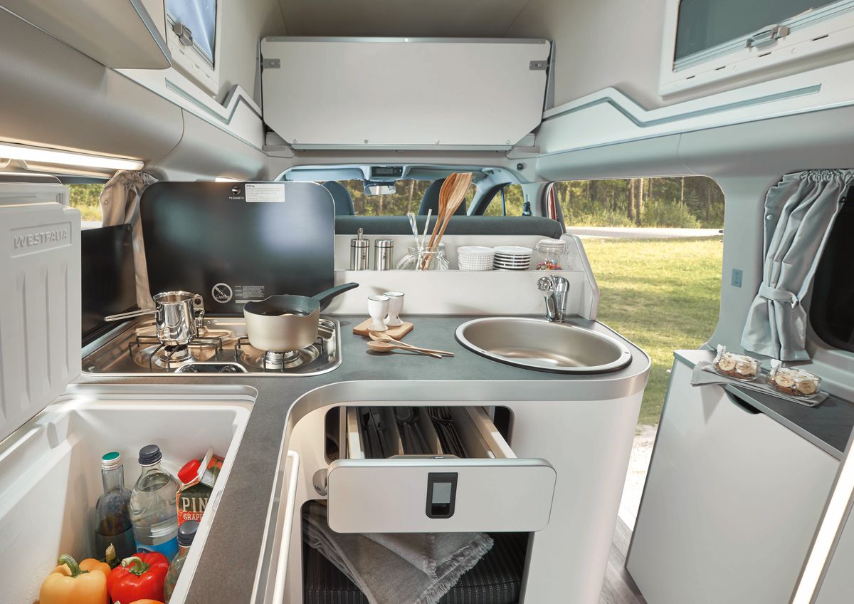 The interior of a minivan has a gray and white kitchen with sink, dual burner stove top, and fridge. 