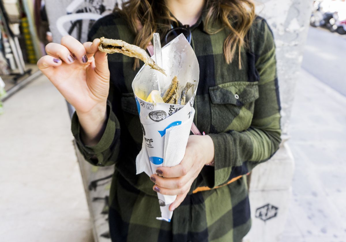 A woman stands on the street holding a piece of fried fish from a paper cone full of fried items
