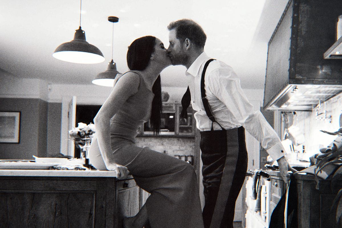 A black-and-white photo of Meghan sitting on a kitchen island and Harry standing and kissing her.