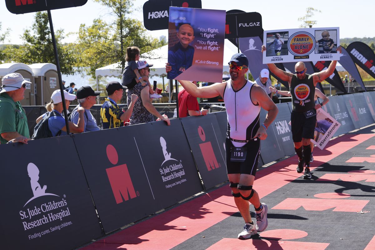 Christopher Corbett and Christopher Frunzi hold up signs to honor their children as they approach the finish line during the IRONMAN 70.3 Memphis on October 1, 2022 in Memphis, Tennessee.