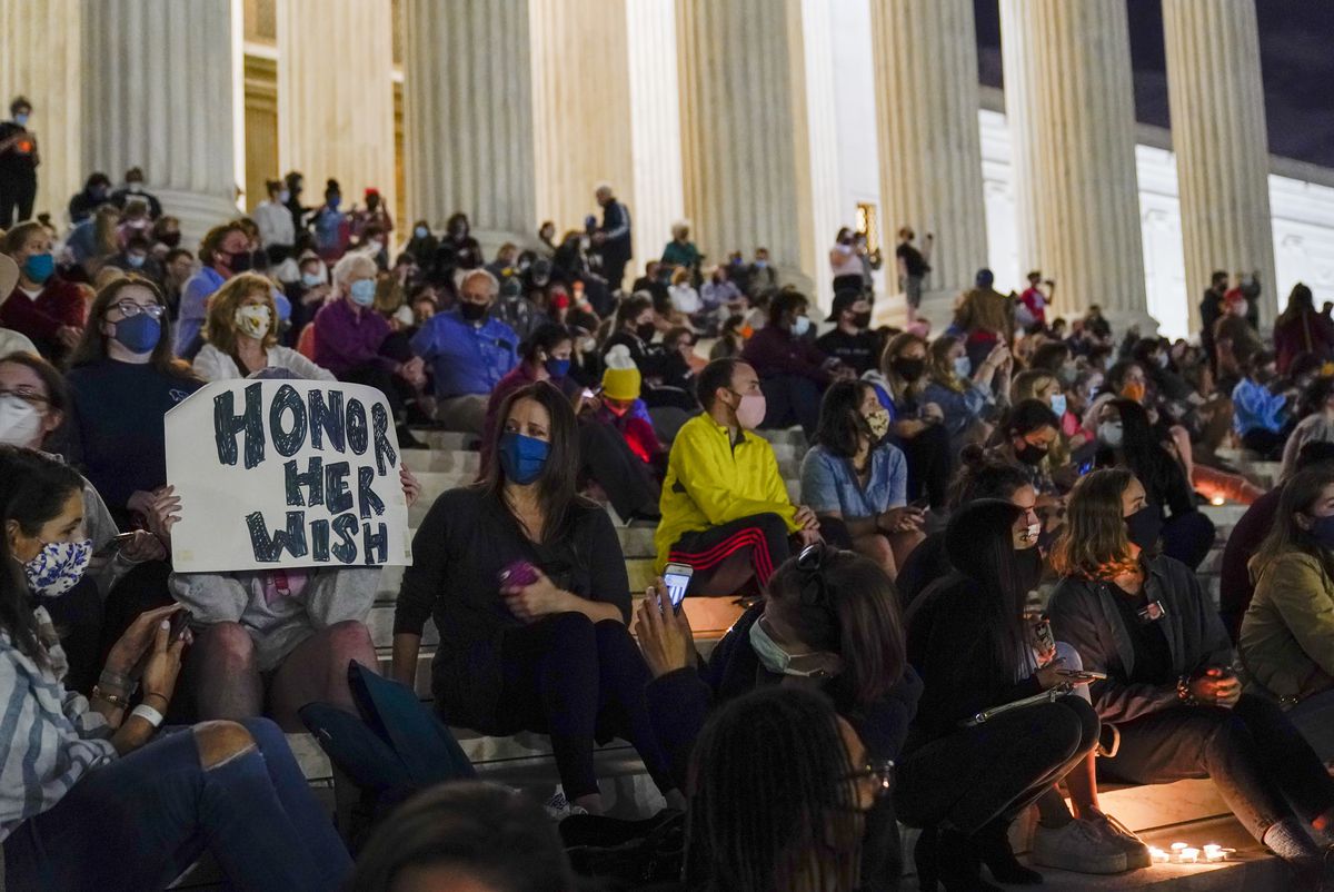 Mourner at the Supreme Court carries a sign saying “Honor Her Wish” on a white posterboard in bold black letters. The mourner is surrounded by a dense, but masked, crowd on the Supreme Court’s steps.