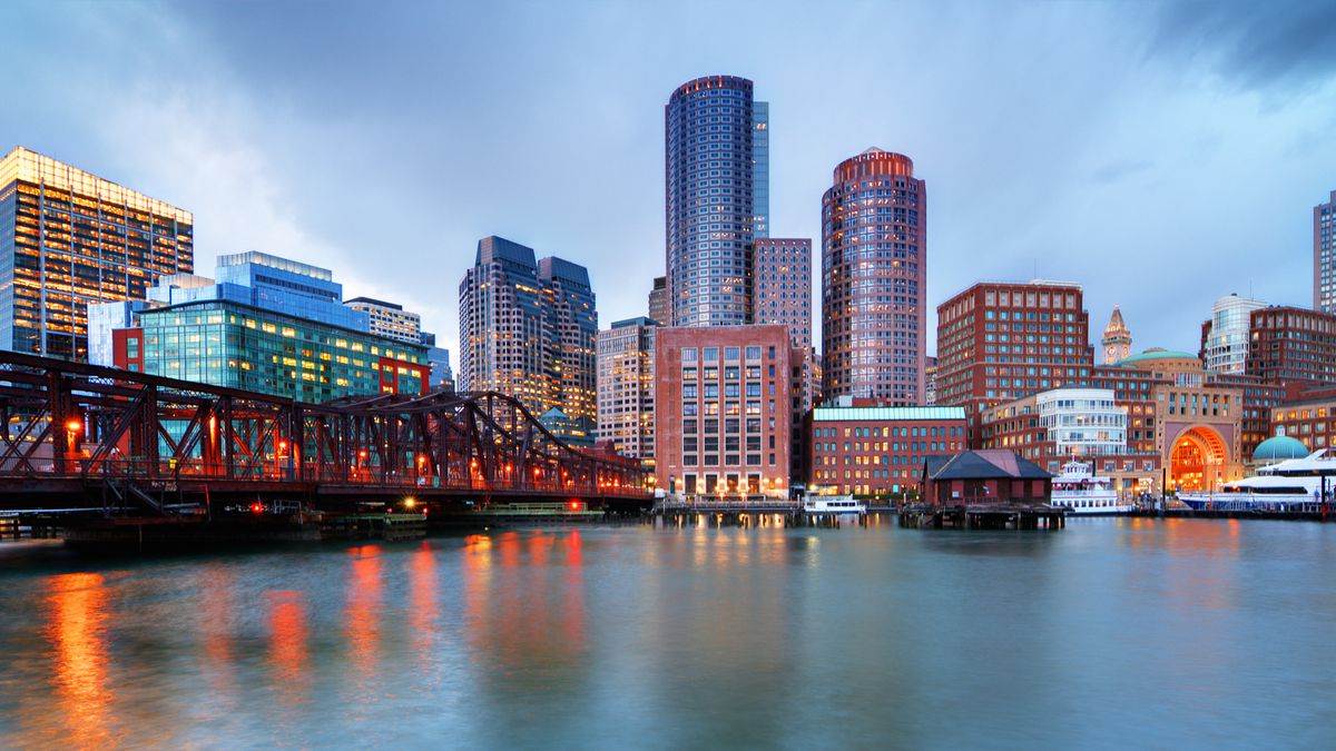 A view of the Boston skyline, featuring Financial District skyscrapers, water, and a cloudy gray sky
