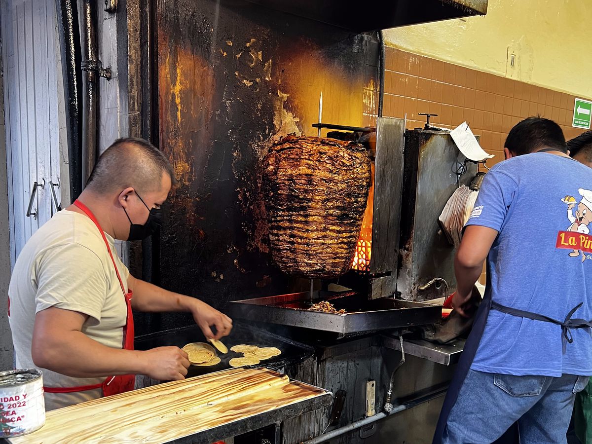 Two cooks work around a trompo, charring from the heat of the fire in an open kitchen.