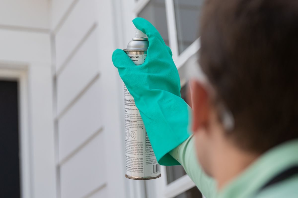 A pest control specialist wearing a green shirt and green gloves sprays pest control solution in the direction of an insect nest on a white house. 