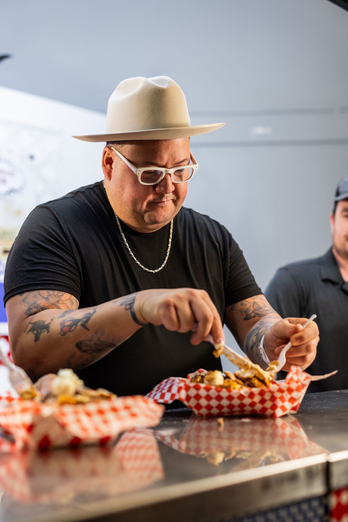 Celebrity chef Graham Elliot looks serious as he uses a fork and knife to eat a dessert at the state fair. Another man is half in the frame to his right, also looking serious.