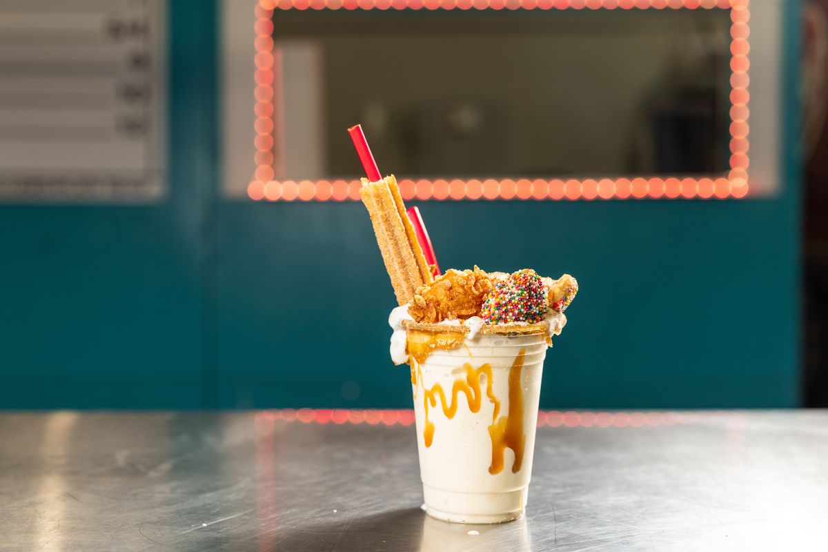 A milkshake sits on a table. It is loaded on top with cookies, fried balls, and the straw is encompassed in a churro, while caramel is smeared on the sides of the clear plastic cup.