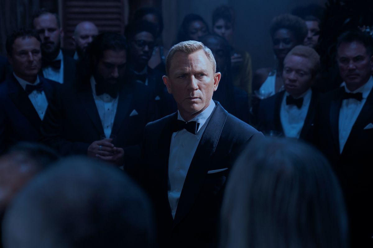 Daniel Craig as James Bond in No Time to Die, standing in a spotlight, surrounded by formal-suited members of SPECTRE