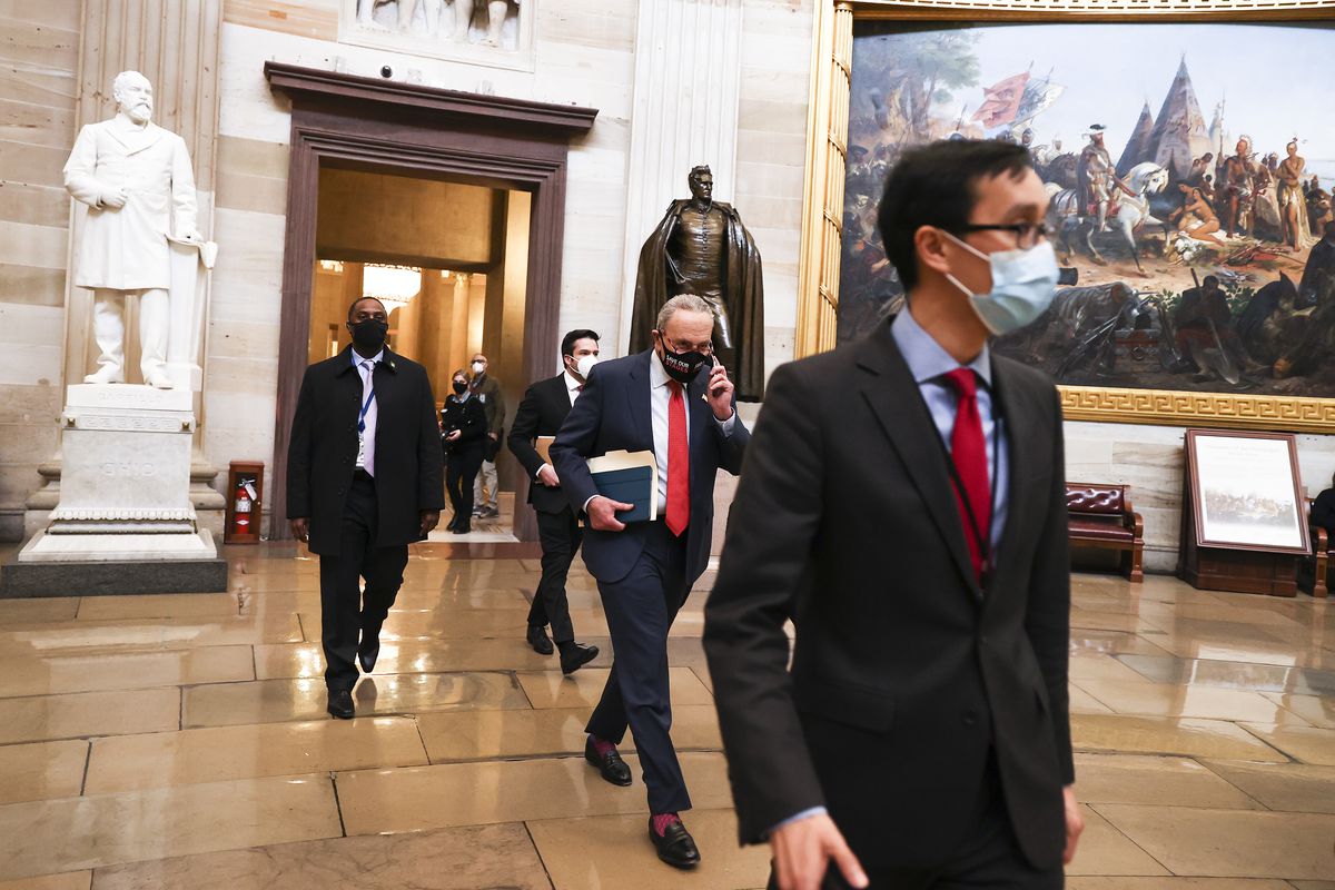 Schumer, in a black mask, dark suit, red tie, black penny loafers, and bright red socks peeking out from under his slacks, walks briskly through the Capitol rotunda, dark oil paintings and large statues of men looking skyward behind him. He is surrounded by aides in suits, and by a large masked man with a shaved head who appears to be a guard.