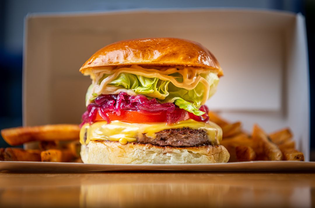 A tall burger topped with melted yellow cheese, sliced red tomatoes, pale green lettuce, and rings of purple onions on a golden bun. Fries in the background.