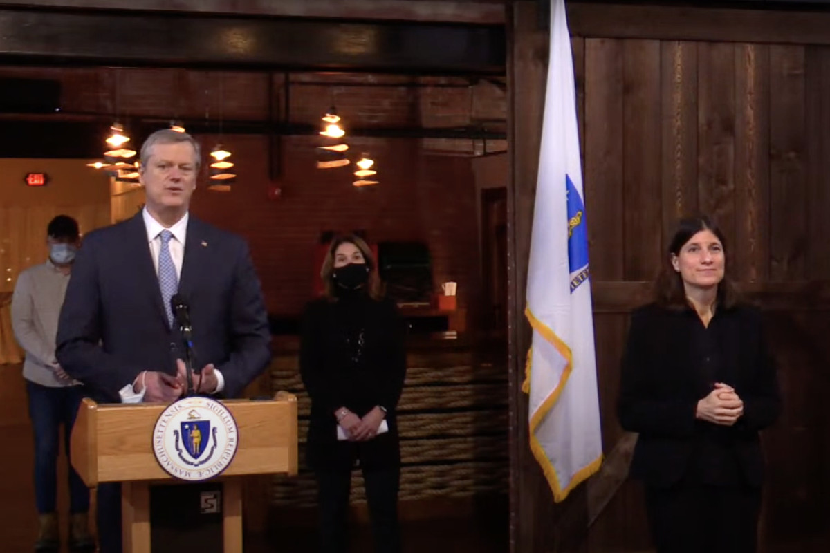 Mass. Gov. Charlie Baker stands at a podium. An American Sign Language translator stands to his left (in the right of the frame), and Lt. Gov. Karyn Polito stands behind him. 