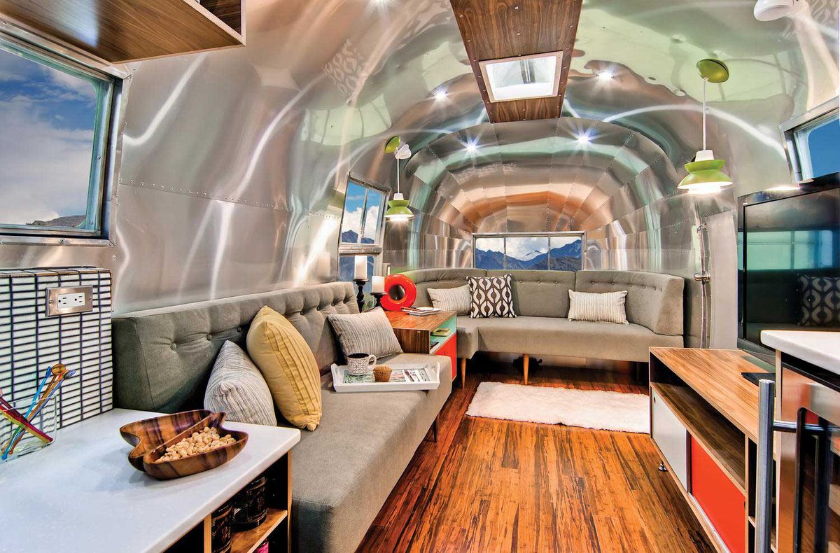 Interior of renovated airstream, with a green midcentury-inspired couch, wood floors, and gleaming aluminum interiors. 