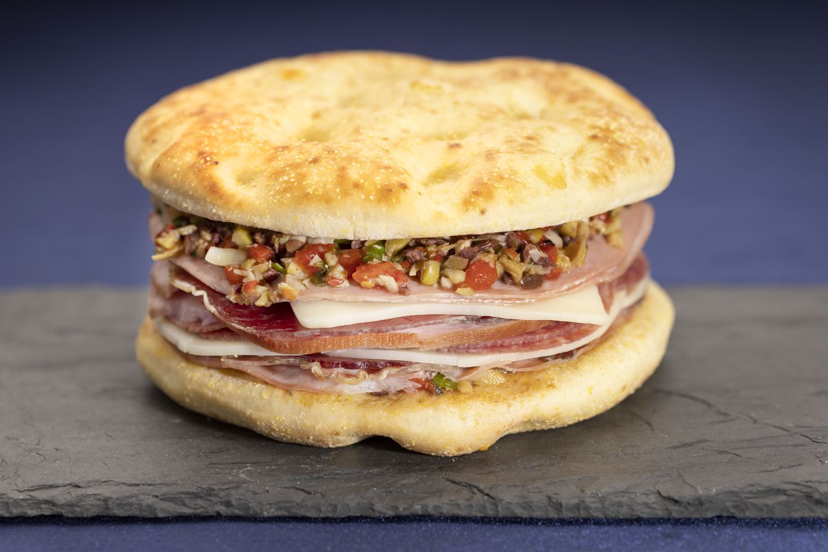 Focaccia bread is topped with house-made olive salad and layers of capricola, mortadella, ham, salami, provolone, and mozzarella cheeses.&nbsp;&nbsp;