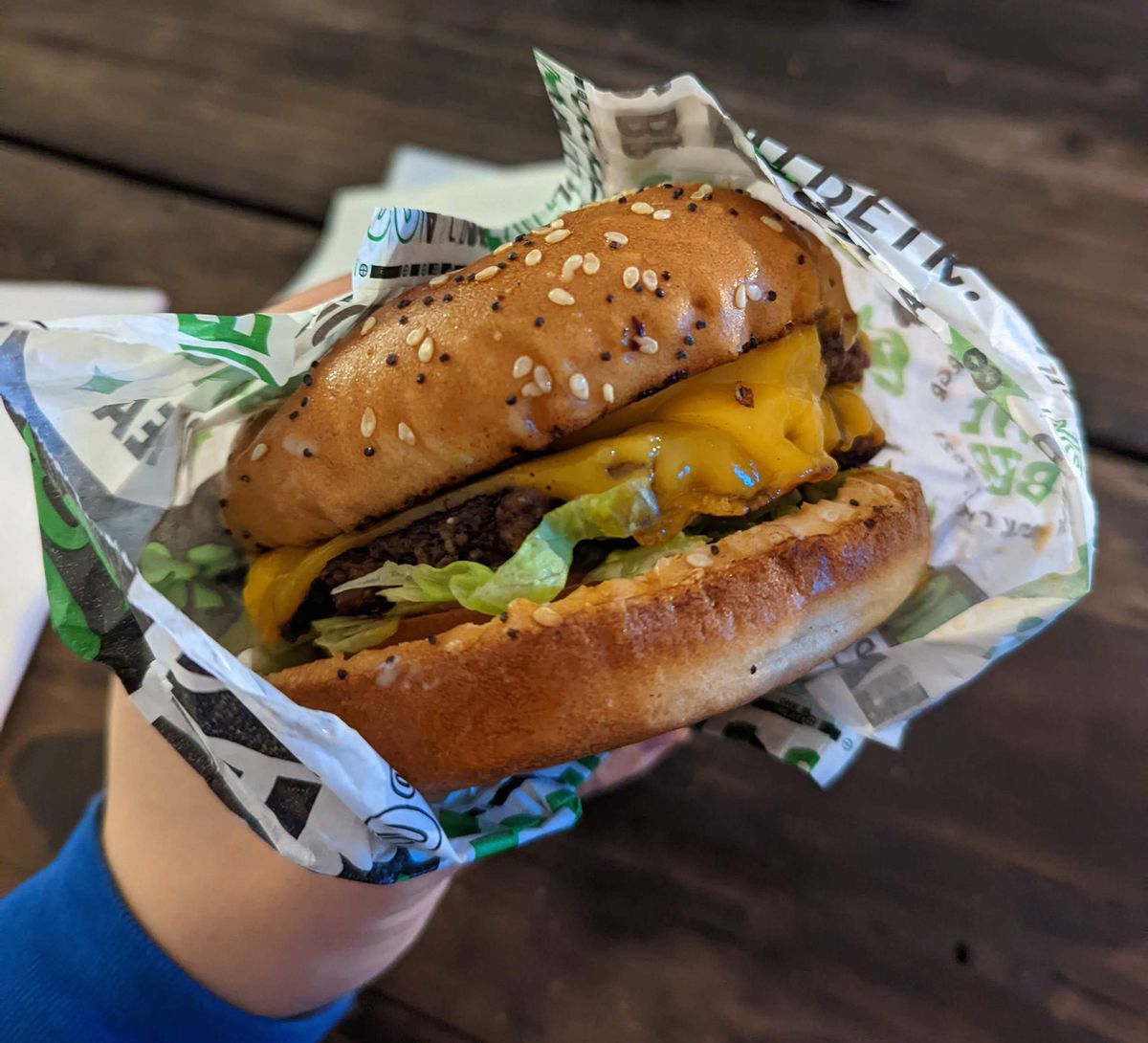 A hand clutches a cheesy smash burger that’s partially wrapped in checkered parchment paper.