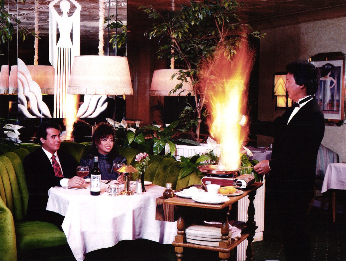 A vintage photograph of diners sitting in a round green booth watching a waiter flambe a dish at the table.