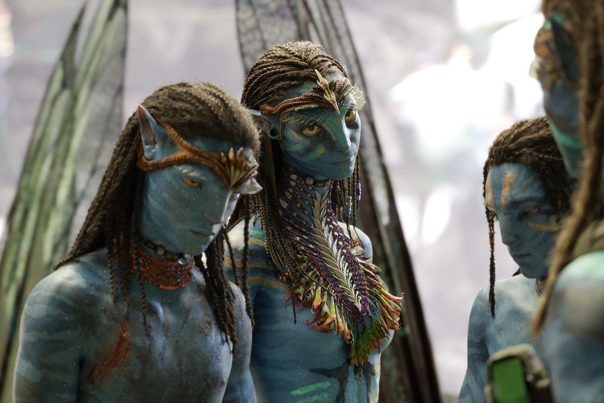 a blue-skinned na’vi boy bows his head in avatar: the way of water. next to him, his mom gives an annoyed look at her husband