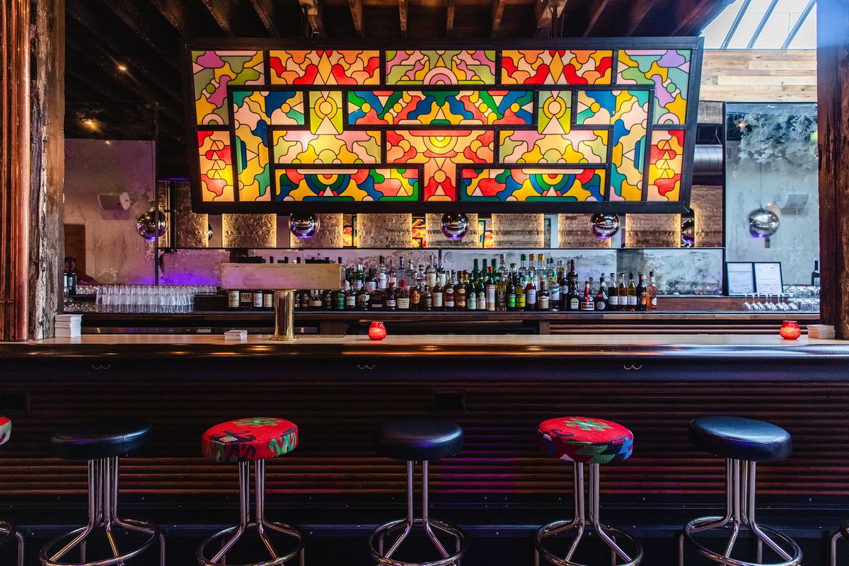 A bar with a large piece of stained glass with a colorful, geometric design overhead and a row of stools in the foreground