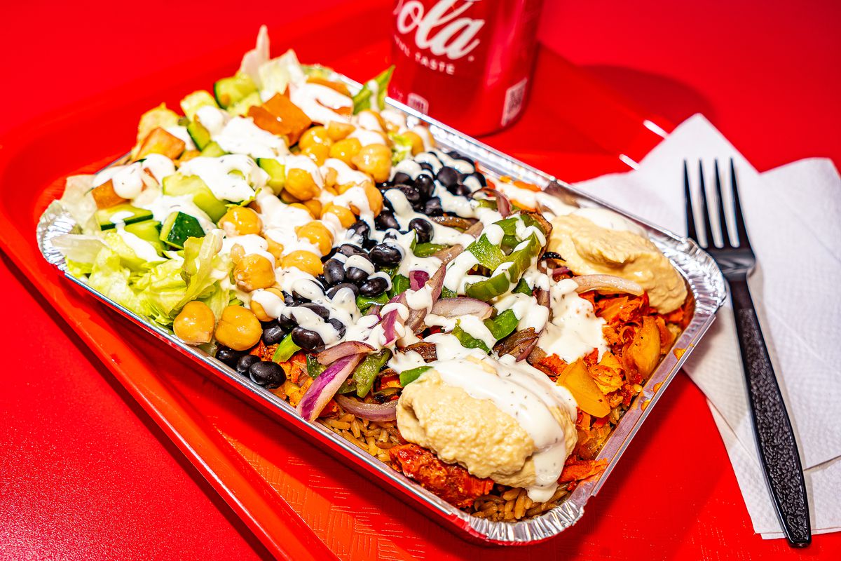 An aluminum container of chicken and rice, topped with white sauce, sits on a bright red tray on a bright red table. A plastic fork and can of Coca-Cola are visible in the background.