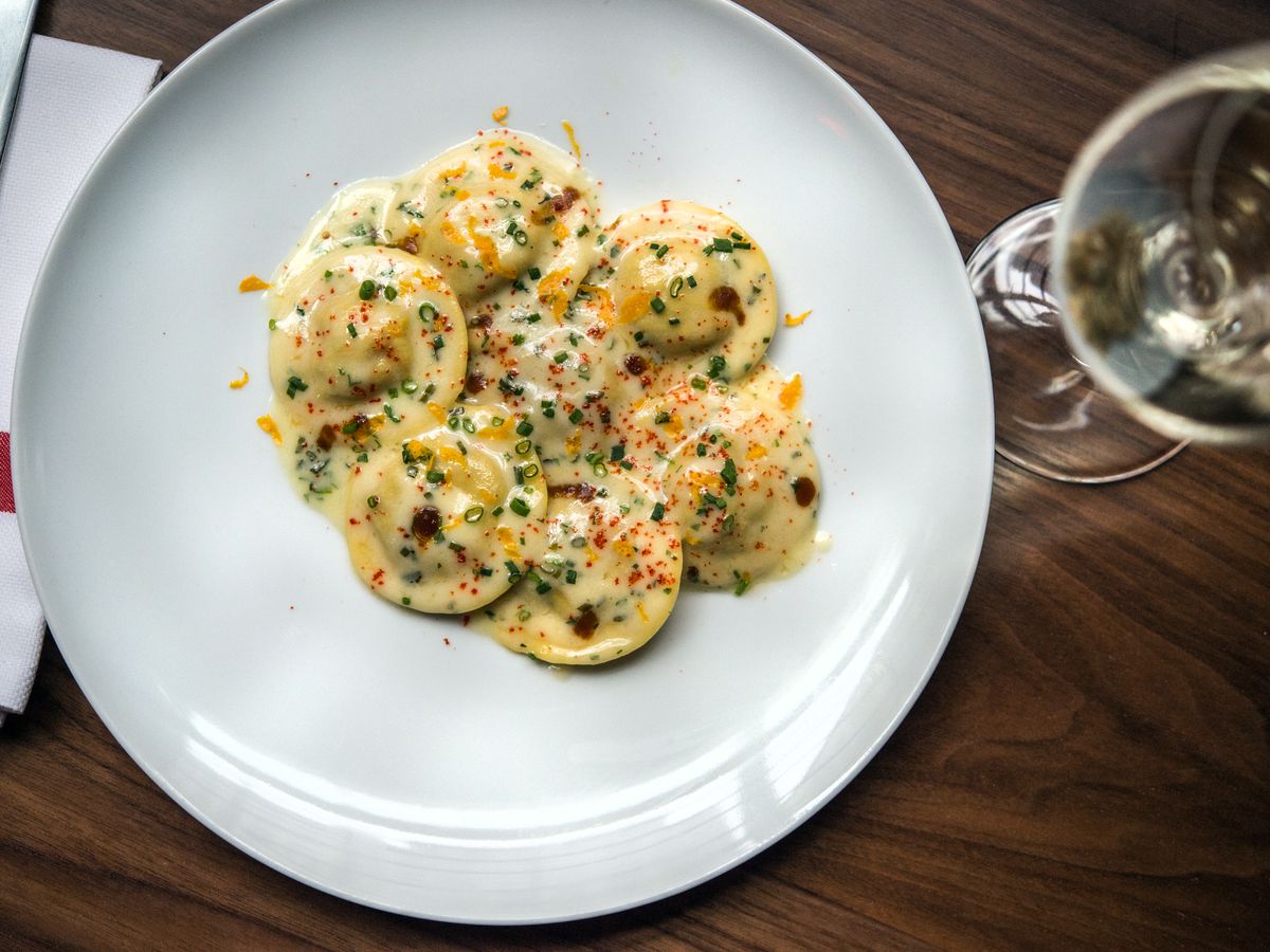 A saucy pile of lobster ravioli on a white plate with a wine glass to the right.