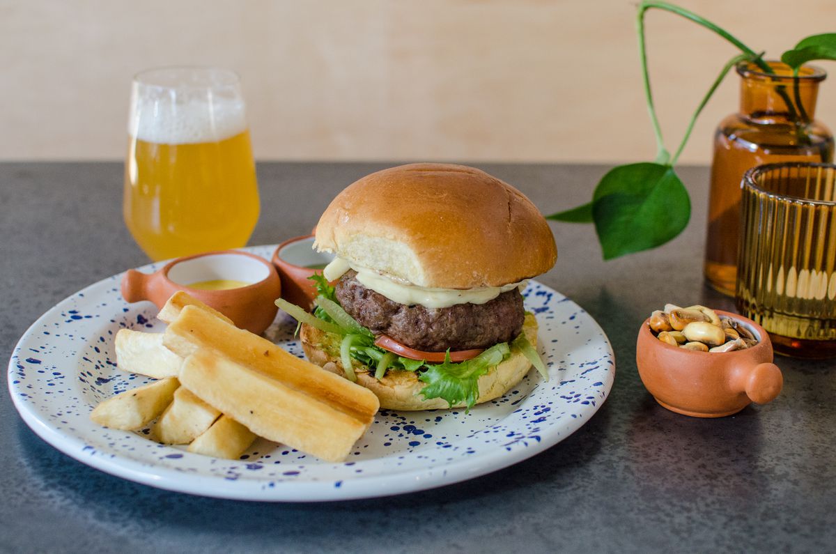 A cheeseburger sits on a white plate with dark blue speckles. The burger is accompanied by thick fries, and there’s a glass of beer in the background.
