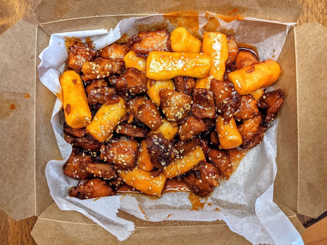 Overhead view of a cardboard takeout container full of small pieces of chicken and tube-shaped Korean rice cakes coated in a reddish brown sauce
