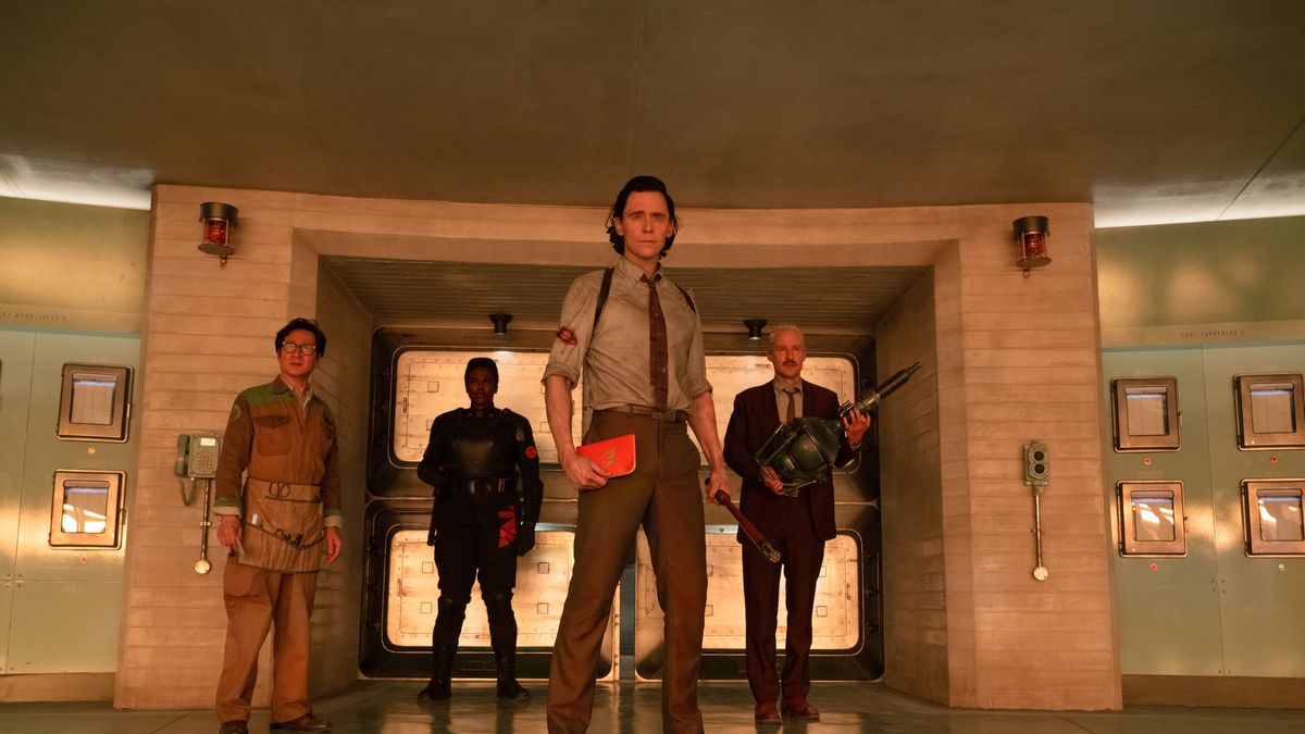 Loki stands with an orange handbook in his hand with the members of the TVA, including Agent Mobius and Ouroboros, standing behind him.