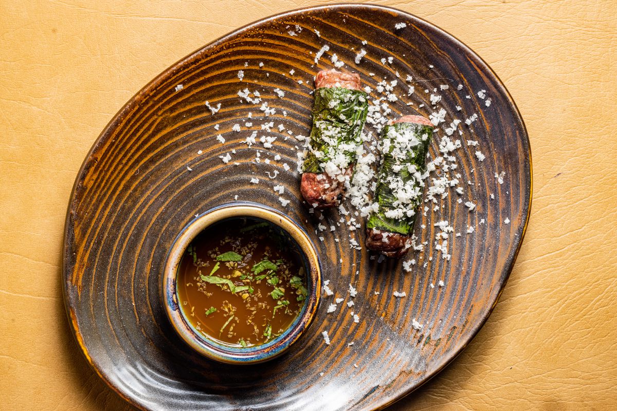 On a wood-looking plate, two wagyu sausages are rolled in pepper leaf and served with a dipping sauce.