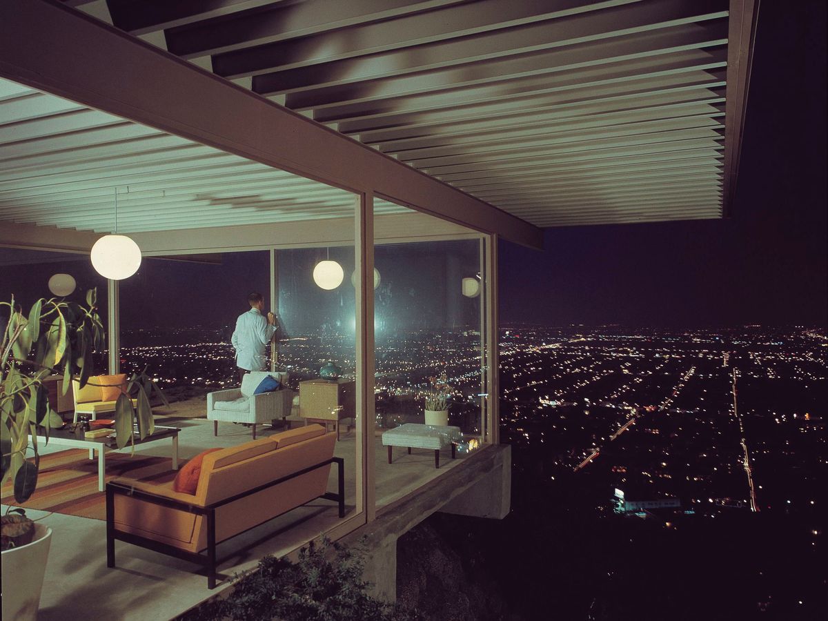 A corner of the Stahl house in Los Angeles. There are floor to ceiling windows in a room with an orange couch and round light fixtures. There is a person looking out of the window at a cityscape. It is night. 
