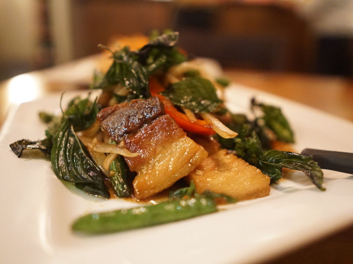 A picture of hunks of pork belly tossed with greens and snap peas, served on a white plate.