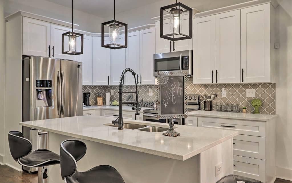 White kitchen with gray tile backsplash, island with two barstools, and pendant lights. 