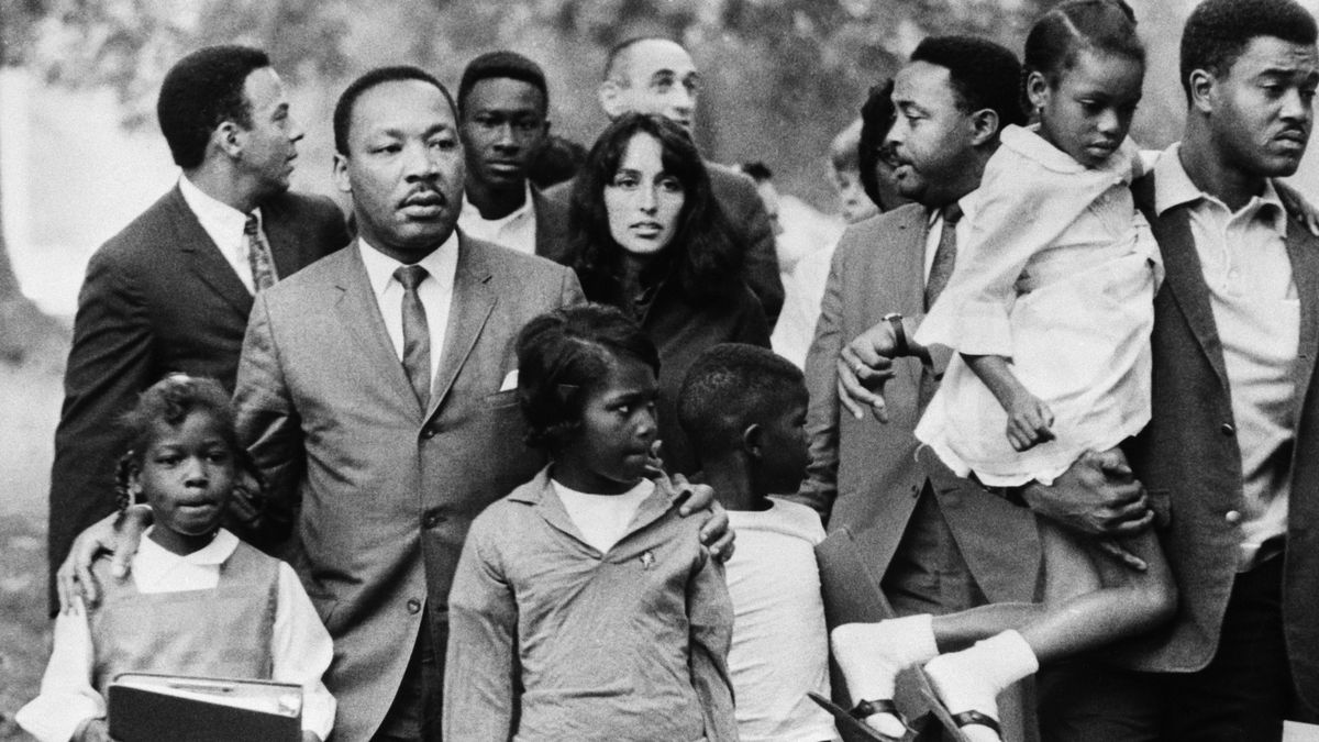An archival black and white photograph of Dr. Martin Luther King escorting a group of Black children to their newly integrated school, joined by Joan Baez, Andy Young, and Hosea Williams, among others.
