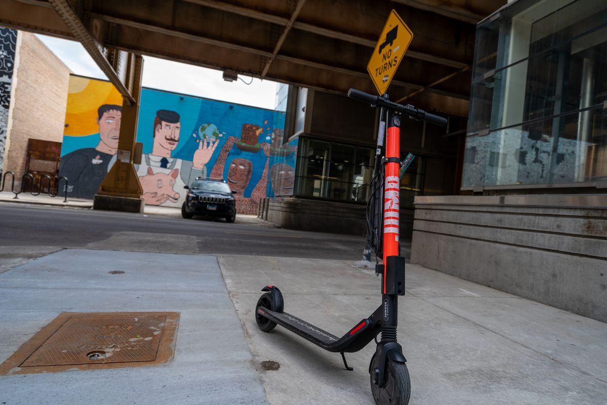 A Jump e-scooter parked on the sidewalk corner near L tracks with a colorful mural in the background