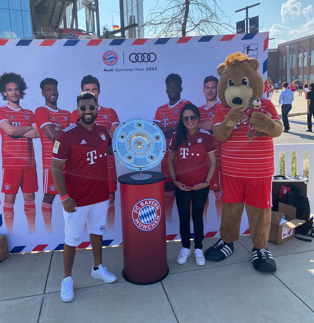 Mr. and Mrs. Soundz pose with Berni the bear and the Meisterschale in front of a poster featuring the Bayern players.