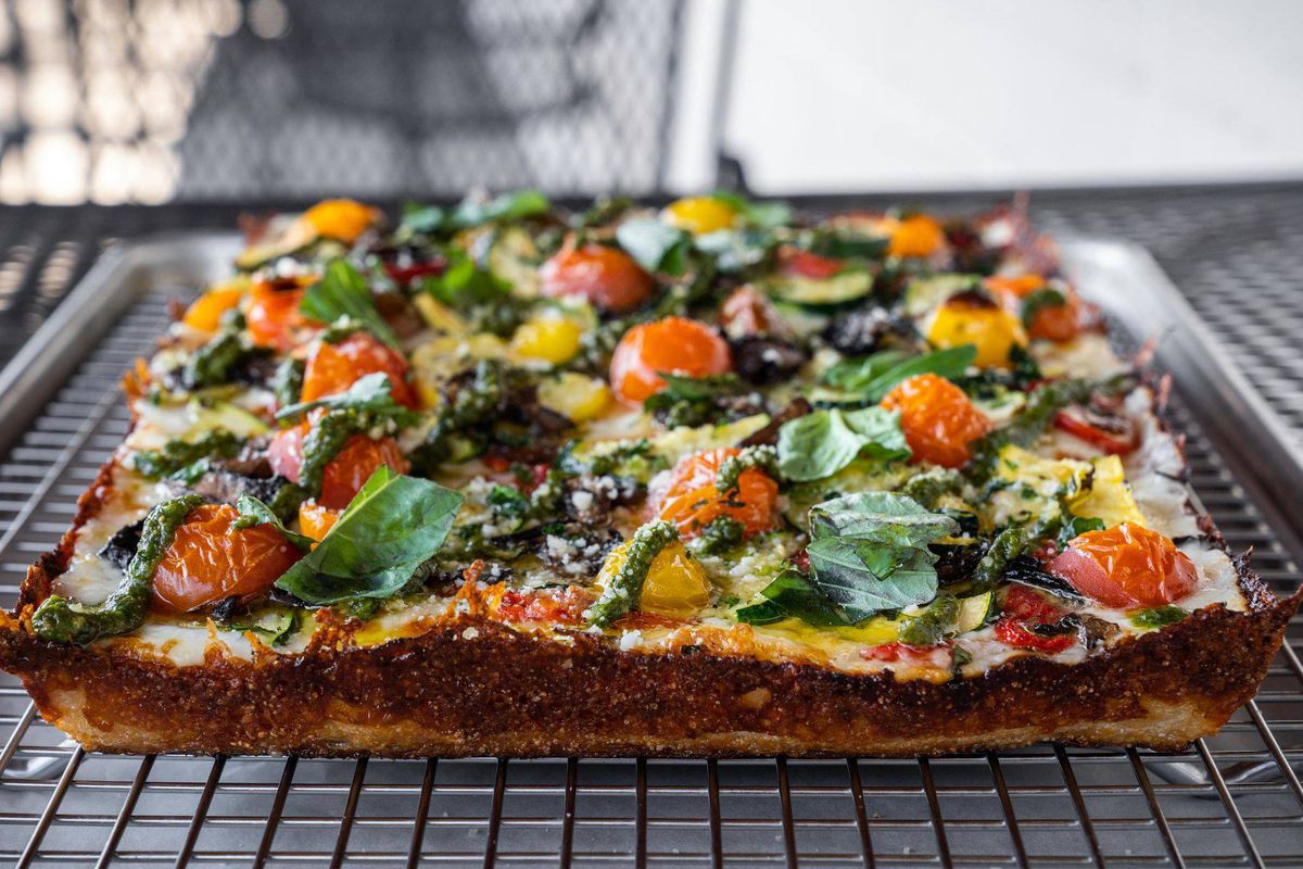 a Detroit-style pizza with crispy edges cooling on a wire rack. The pizza is topped with red and yellow cherry tomatoes and spinach